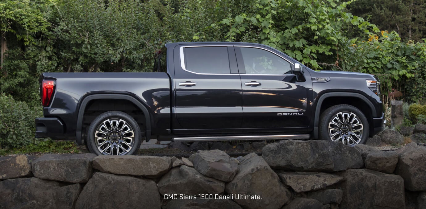 Side profile view of a Titanium 2026 GMC Sierra 1500 Denali Ultimate parked behind a low stone.