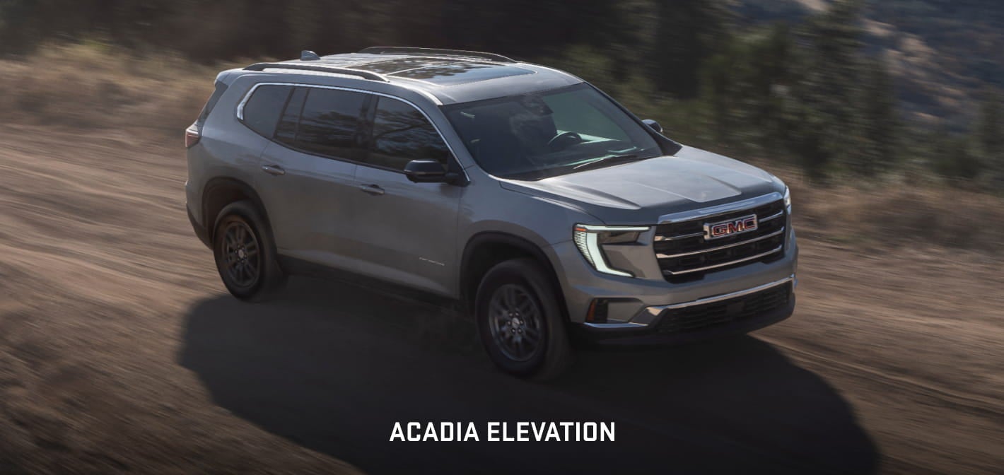 Gray GMC Acadia Elevation driving on an unpaved road, shown from a front three-quarter angle.