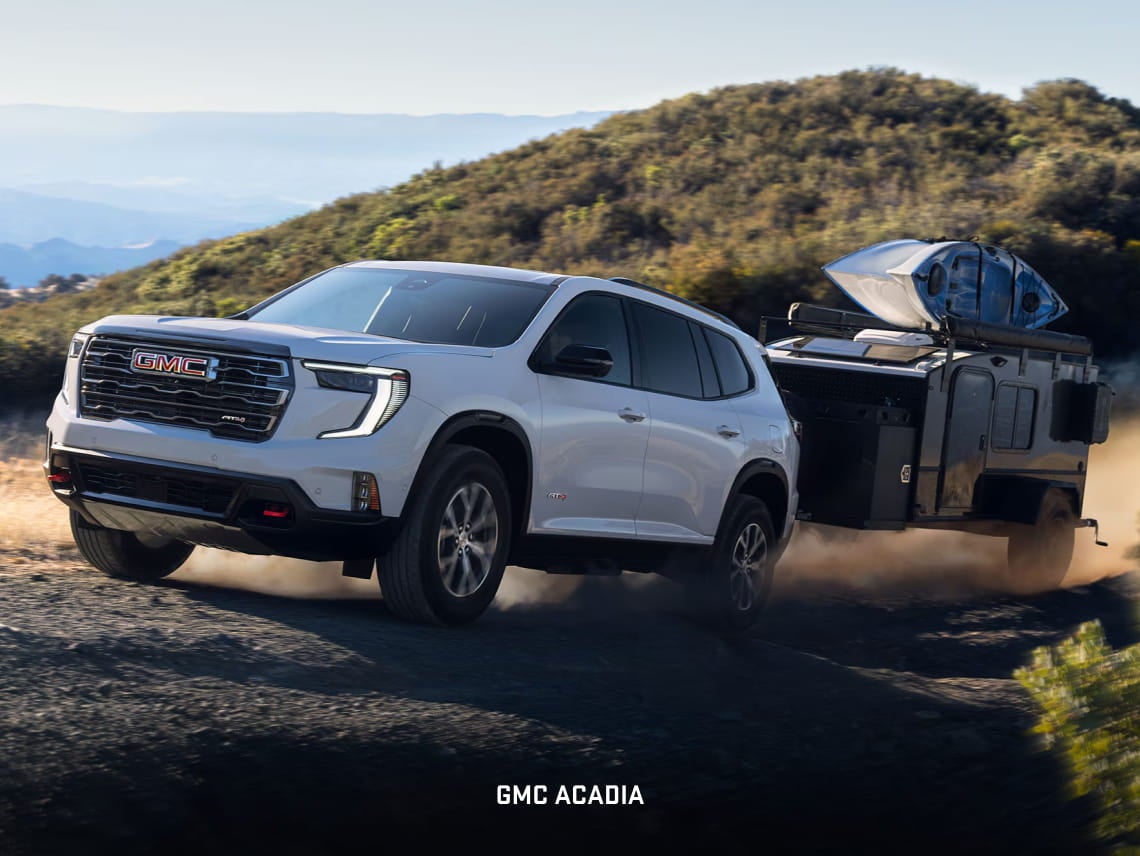 White GMC Acadia towing an adventure trailer with a kayak on an unpaved road.
